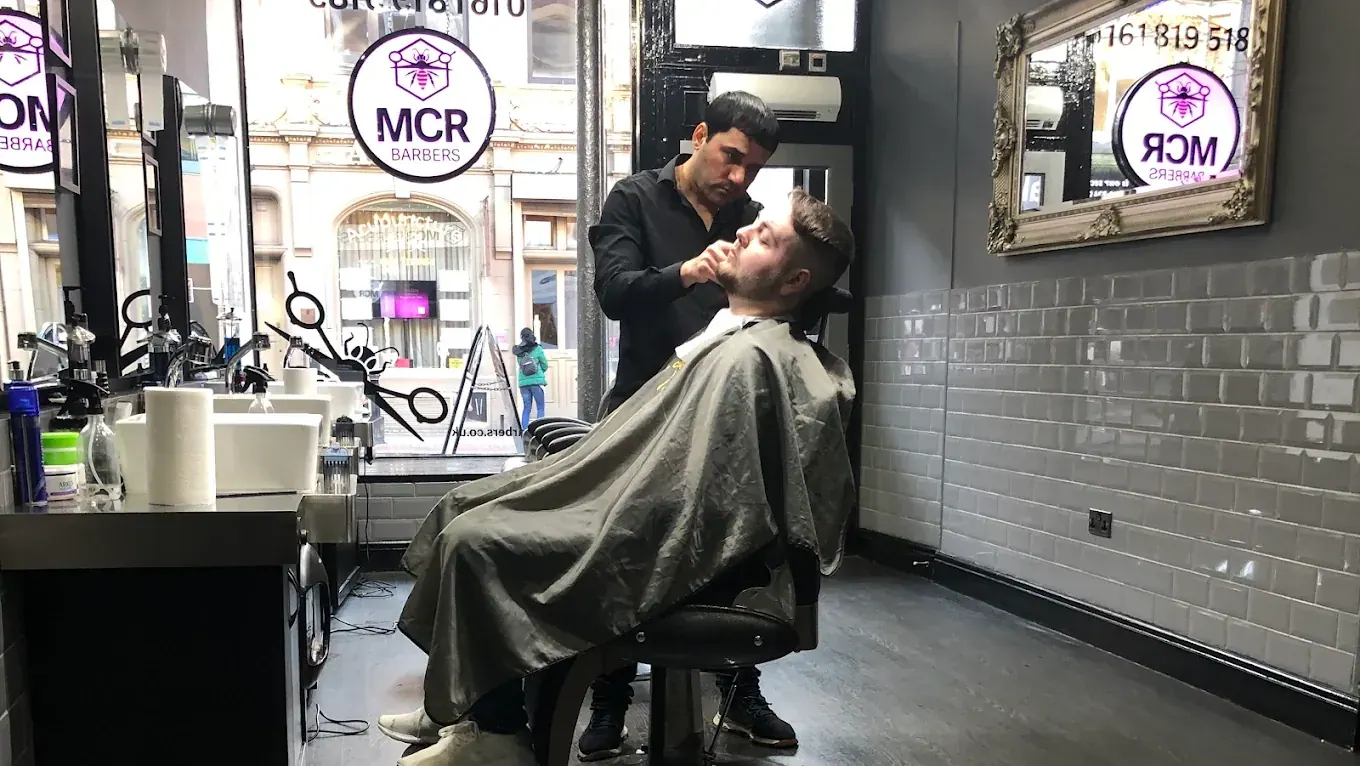 A barber shaping a client's beard at the front window, under the MCR Barbers sign