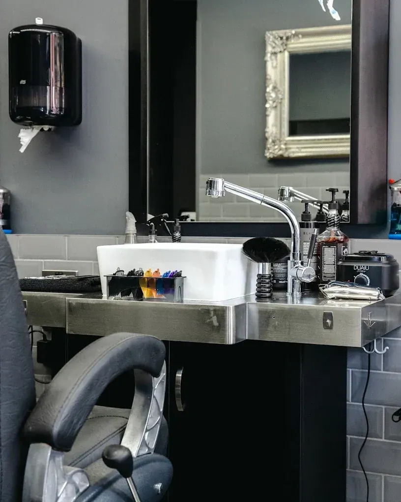 A barber station close up: white basin, chrome tap, combs and a neck brush, with a silver-framed mirror behind